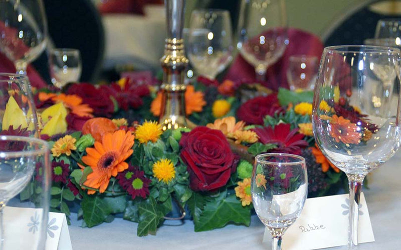 Stor blomsterkrans til bordet. Bundet med røde roser kombineret med orange og gule blomster. Kransen ligger på et rundt bord omsluttende en fem-armet lysestage